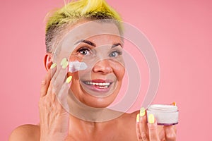 Mature woman with stylish yellow dyed hair and bare shoulders applying anti-aging cream on pink studio background