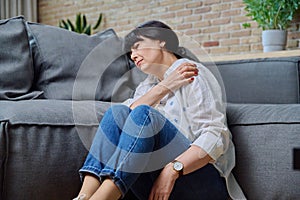 Mature woman suffering from pain in her shoulder, sitting at home on floor