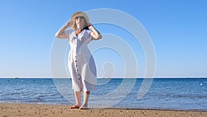 mature woman in sunglasses in a straw hat and white dress walks along the blue sea coast on a sunny summer day, enjoying