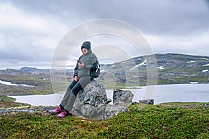 Mature woman takes pause and rests on stone, looking at her mobile, hiking high in Norwegian mountains. Healthy lifestyle. Norway