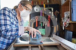 Mature Woman Upcycling Furniture In Workshop At Home Using Electric Sander