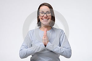 Mature woman wearing glasses keeps palms together, has pleased expression