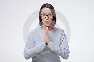 Mature woman wearing glasses keeps palms together, has pleased expression