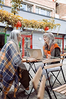 Mature women business partners with laptop discuss project on outdoors cafe terrace