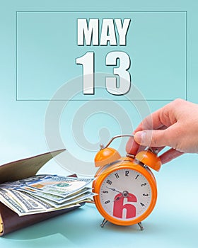 May 13th. Hand holding an orange alarm clock, a wallet with cash and a calendar date. Day 13 of month.