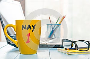 May 1st. Day 1 of month, calendar on morning coffee cup, business office background, workplace with laptop and glasses