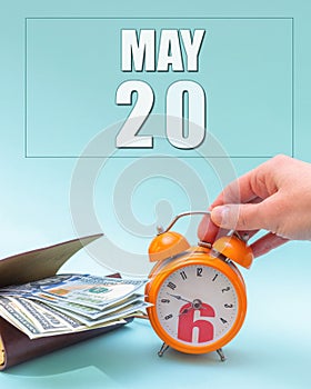 May 20th. Hand holding an orange alarm clock, a wallet with cash and a calendar date. Day 20 of month.