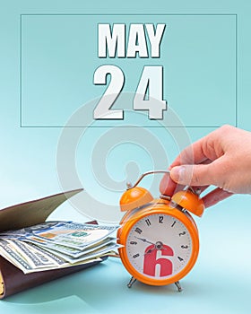 May 24th. Hand holding an orange alarm clock, a wallet with cash and a calendar date. Day 24 of month.