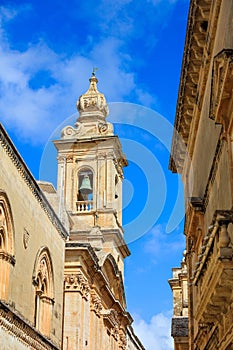 Mdina, Malta. Narrow street wth church belfry and buildings stone facades on blue sky background