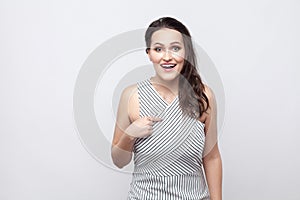 Is it me? Portrait of amazed beautiful young brunette woman with striped dress standing, poining herself and looking at camera