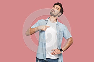 This is me. Portrait of proud haughty handsome bearded young man in blue casual style shirt standing, looking away and pointing photo