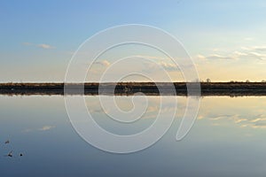 Meadow bank of the river, going beyond the horizon, divide the photo in half. Reflection of the sky in the water of the river. Sun