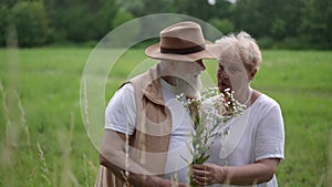 In the meadow, an elderly gray-bearded man in a brimmed hat gives an elderly woman meadow flowers. An elderly couple is