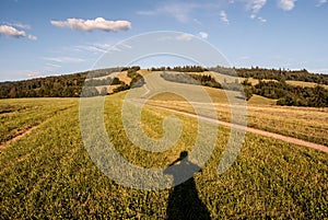 Meadow with pathway, photographer shadow, hill on the background and blue sky with clouds