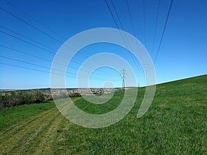 Meadow on a slope above which towers of power lines and in the distance is a village
