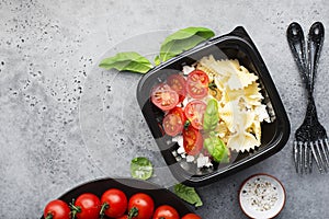 Meal prep. Black lunch boxes. Pasta bows, cherry tomatoes, young cheese, salt, pepper, basil. On a dark background Top