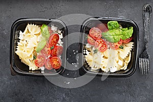 Meal prep. Black lunch boxes. Pasta bows, cherry tomatoes, young cheese, salt, pepper, basil. On a dark background. Top