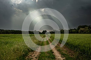 Dirt road through green field and dark stormy sky with clouds