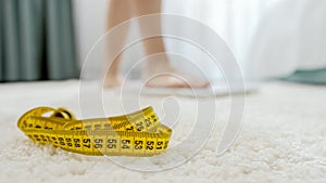 Measuring tape lying on floor next to woman standing on scales in bedroom. Concept of dieting, loosing weight and