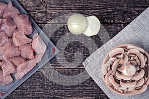 Meat cut into pieces, peeled onion and a bowl of chopped champignons on an old wood texture background.