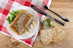 Meat jelly, basil, slices of garlic in white plate, knife and fork, pepper shaker on napkin, slices of bread on wooden table. Top