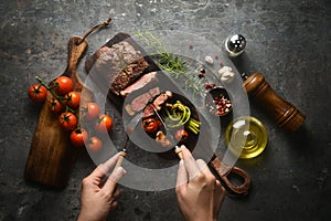 Meat steak serving on wooden butcher board with various ingredients surrounding, and hands holding fork and knife to cut a pieces