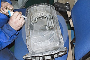 A mechanic in a blue uniform applies a gray sealant from a tube with his hand to the surface of the pallet of an automobile engine