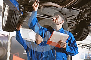 Mechanic in blue work wear uniform inspects the car bottom with his assistant. Automobile repairing service.