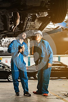 Mechanic in blue work wear uniform inspects the car bottom with a wrench with his assistant. Automobile repairing service.