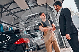 Mechanic with clipboard and man or owner shaking hands at car shop