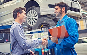Mechanic holding clipboard shaking hands with car owner in the workshop garage. Car auto services concepts