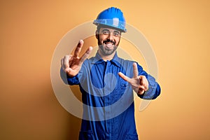 Mechanic man with beard wearing blue uniform and safety helmet over yellow background smiling with tongue out showing fingers of
