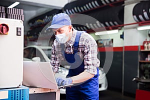 Mechanic man in protective mask with laptop making car diagnostics at service