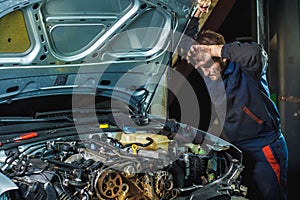 Mechanic standing near a car with an open hood. Tired mechanic looks at the engine, wiping his forehead with his hand.