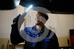 Mechanic, technician, auto engineer checks the underbody of a lifted car with a lamp in the garage of a repair shop. Car warranty