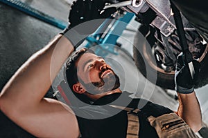 Mechanic in uniform lying down and working under car at auto service garage