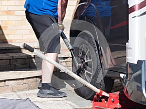 A mechanic uses a metal pole on a wrench to remove the wheel nuts from a motorhome wheel on a private drive. Trolley jack visible