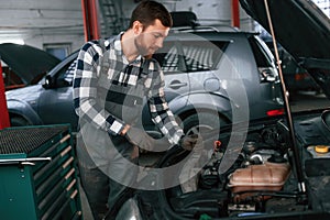 Mechanic is using a tool to repair the car under the hood. Man in uniform is working in the auto salon