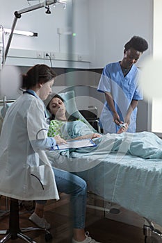 Medic pointing at lab results on clipboard explaining diagnosis to patient in hospital bed