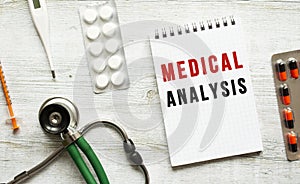 MEDICAL ANALYSIS is written in a notebook on a white table next to pills and a stethoscope