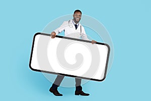 Medical App. Smiling Black Doctor Wearing Uniform Carrying Big Blank Smartphone