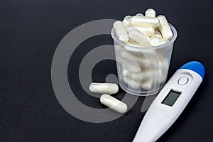 Medical capsules, tablets and clinical thermometer inside measuring cup on black background. Side view