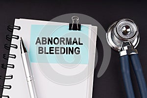 On a black surface, a stethoscope, a notebook, a pen and a blue sticker with the inscription - ABNORMAL BLEEDING photo