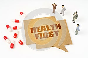 On a white surface are pills, a pen, figurines of people, a sign with the inscription - HEALTH FIRST photo