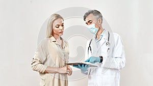 Medical consultation. Mature male doctor in protective mask and young female patient discussing diagnosis, using digital