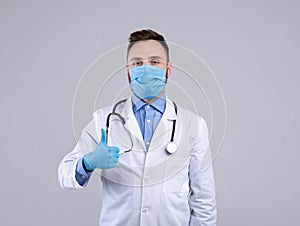 Medical doctor in lab coat, protective mask and rubber gloves showing thumb up gesture over grey studio background
