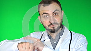 Medical doctor man looking at camera and raises arm to tap on wrist watch. Green screen background