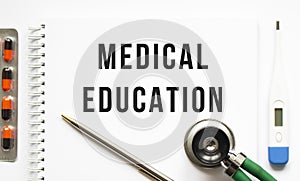 MEDICAL EDUCATION is written in a notebook on a white table next to pills and a stethoscope
