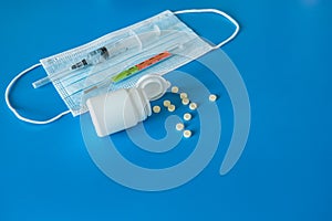 Medical face mask covering mouth and nose, pills, thermometer for measuring body temperature on a blue background.