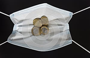 Medical face mask holding money inside of it, on a black background. World coronavirus epidemic and economic losses concept. Coins